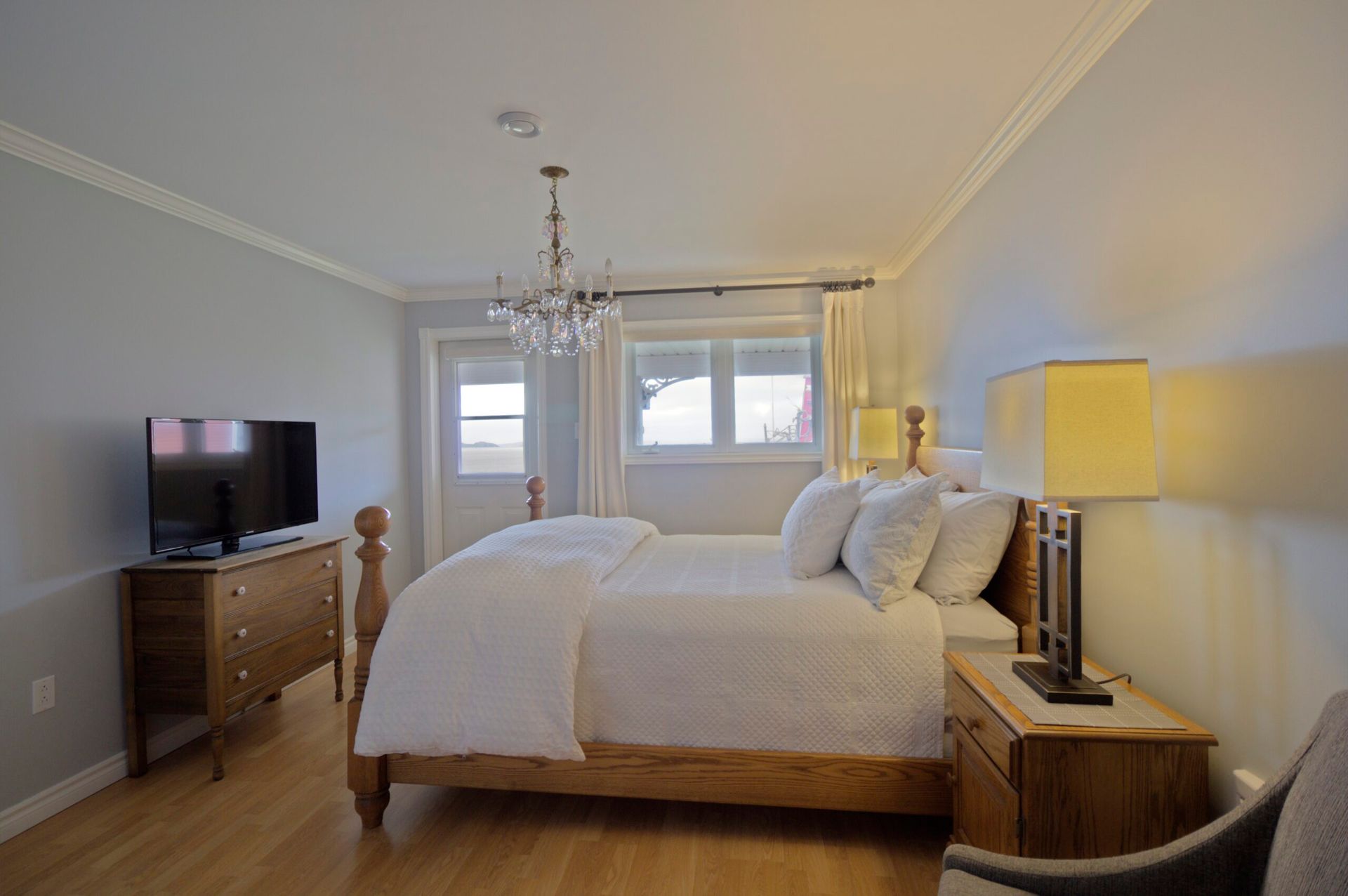 Lichte slaapkamer met een bed, kroonluchter, commode en warme lampen bij een raam.