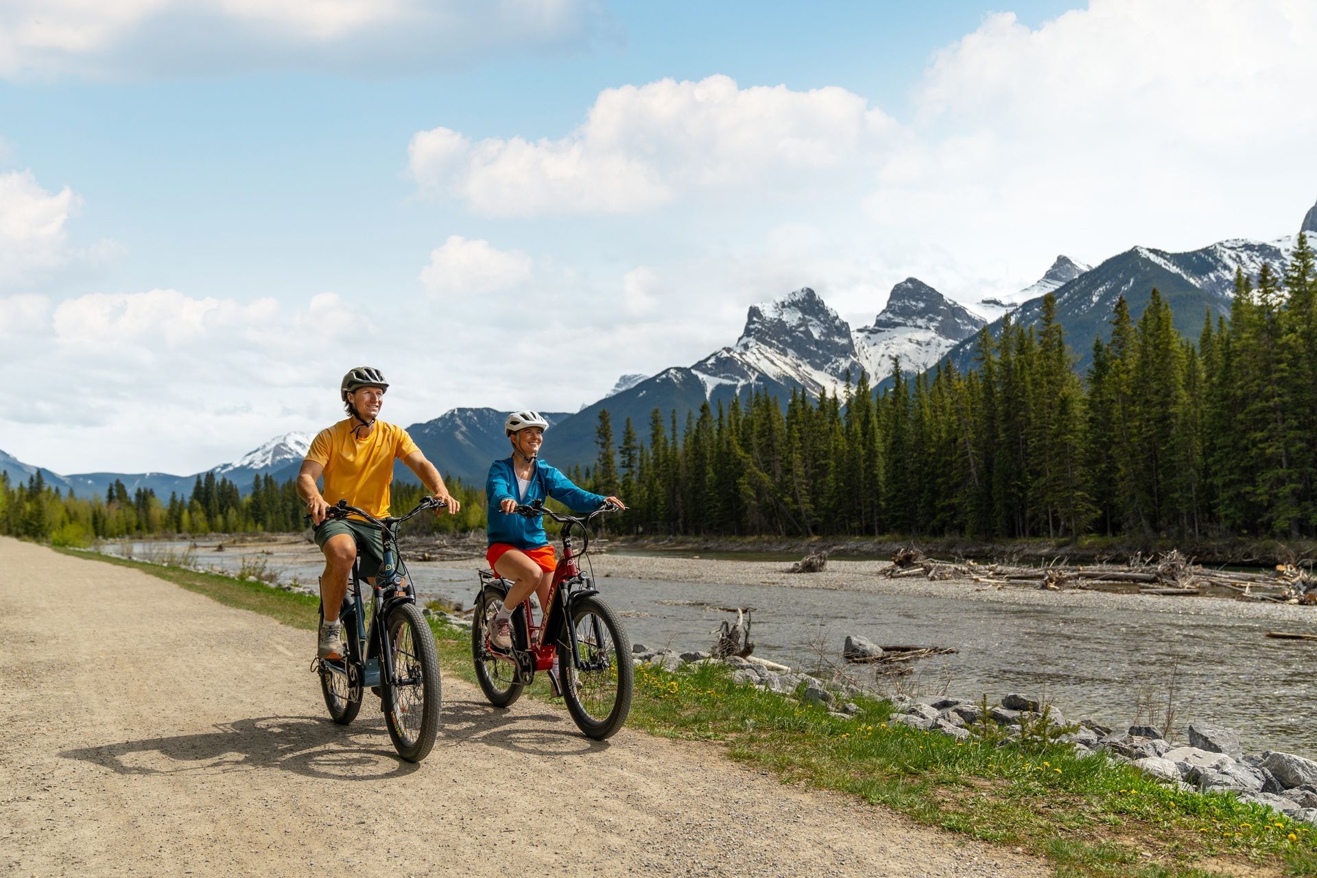 Credits Travel Alberta / ROAM Creative Two cyclists ride along a dirt path with mountains and trees in the background. Sunny, blue sky.
