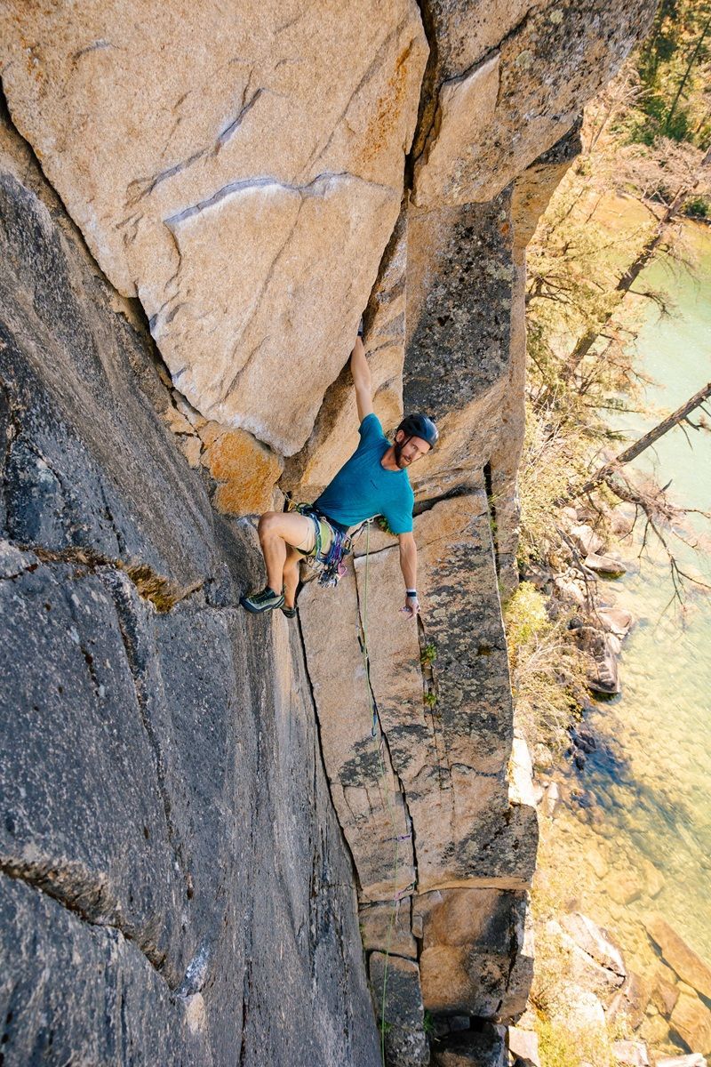 Credits Kai Jacobson Rock climber ascending a steep cliff face; blue shirt, light skin, turquoise water below.