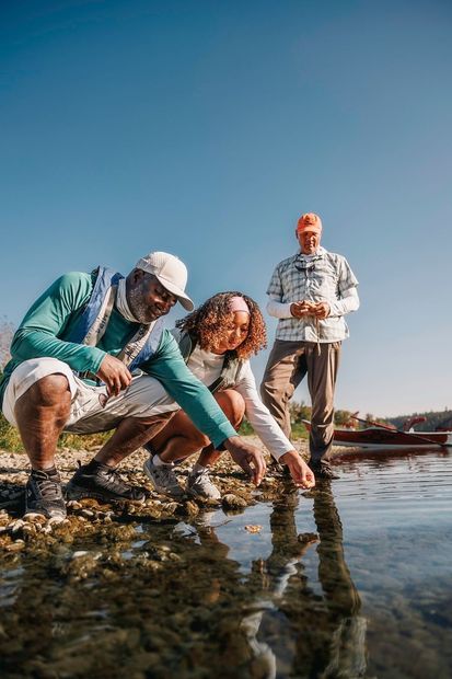@Travel Alberta / Damian Blunt Drie mensen onderzoeken ondiep water. Eén wijst, de anderen kijken toe. Buiten, zonnig weer.
