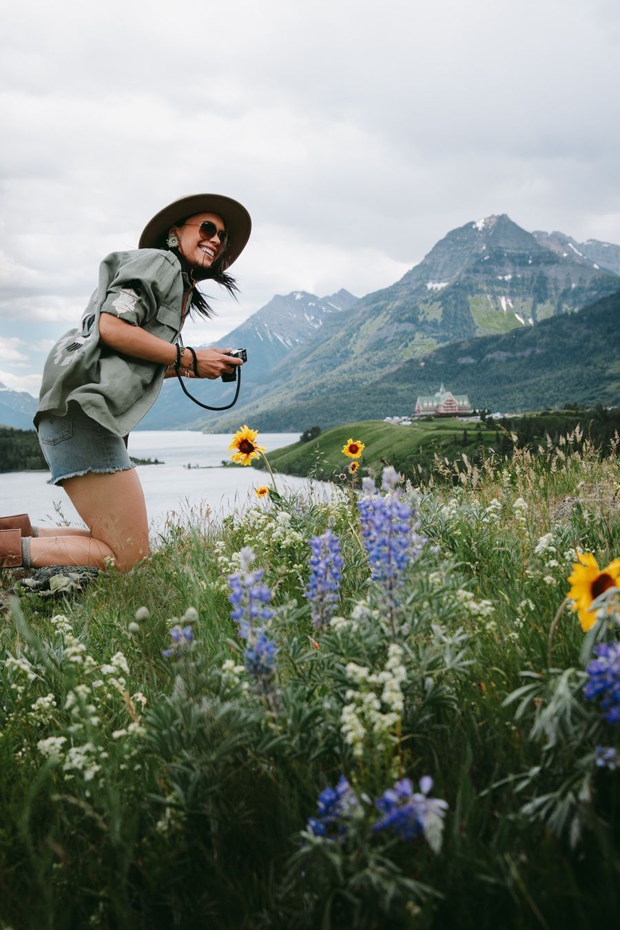 Vrouw knielt tussen de wilde bloemen en fotografeert een meer en bergen.