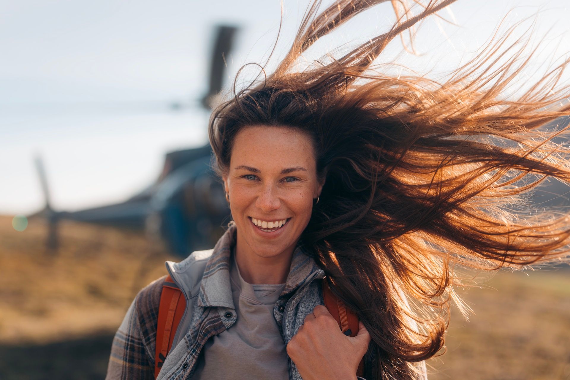 Vrouw met wapperend haar lacht in de buurt van een helikopter.