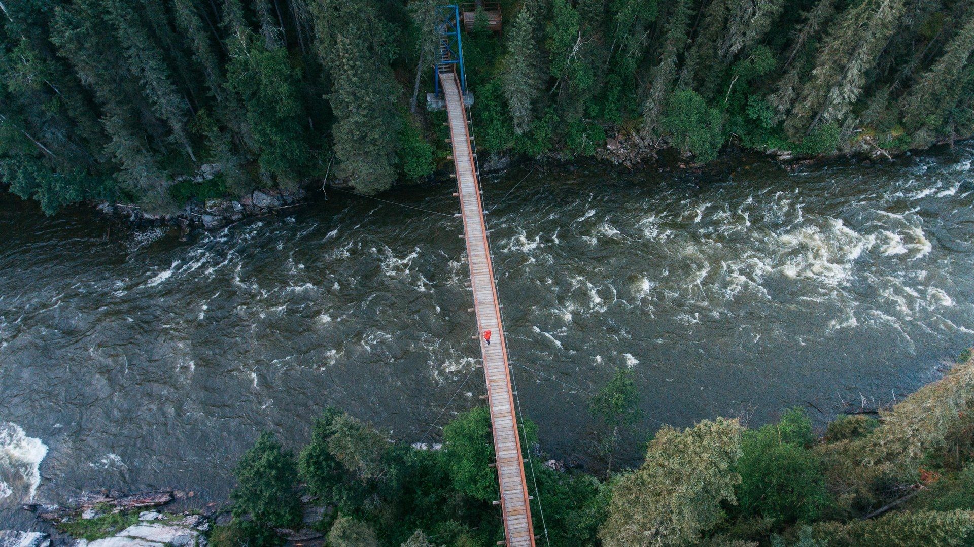 @ Travel Manitoba Overhead view of a wooden suspension bridge over a rushing river in a forest, trees surround.