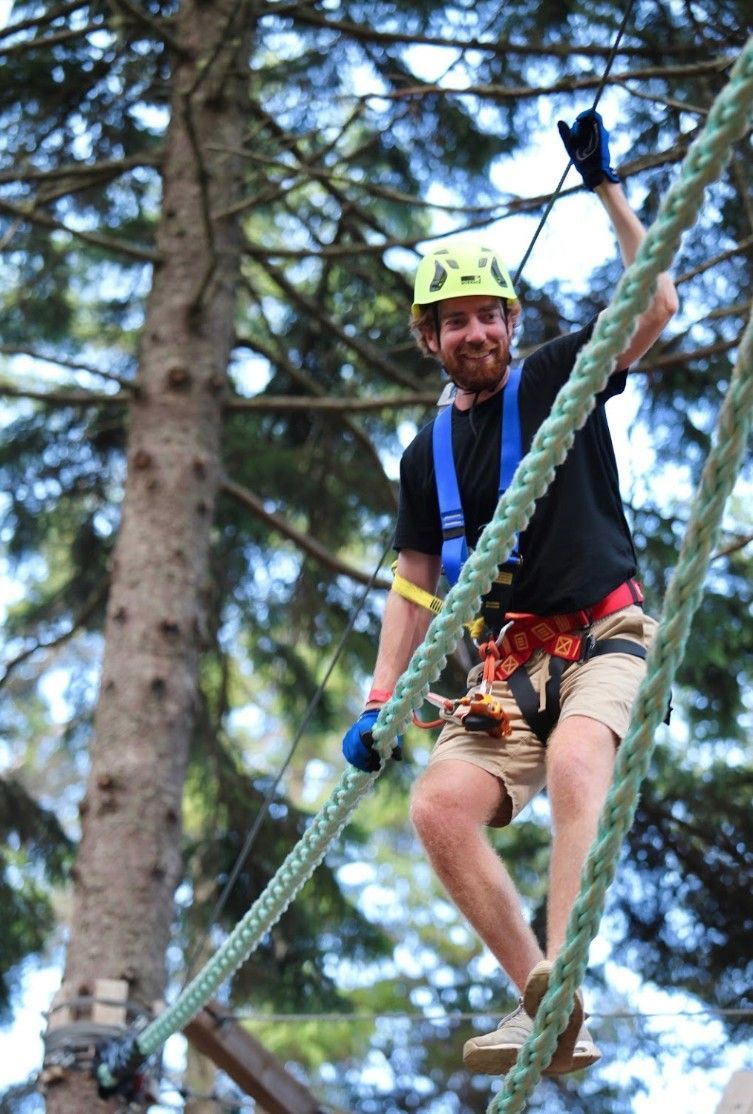 Man on a ropes course, smiling and waving, wearing a helmet and safety harness in a forest setting.