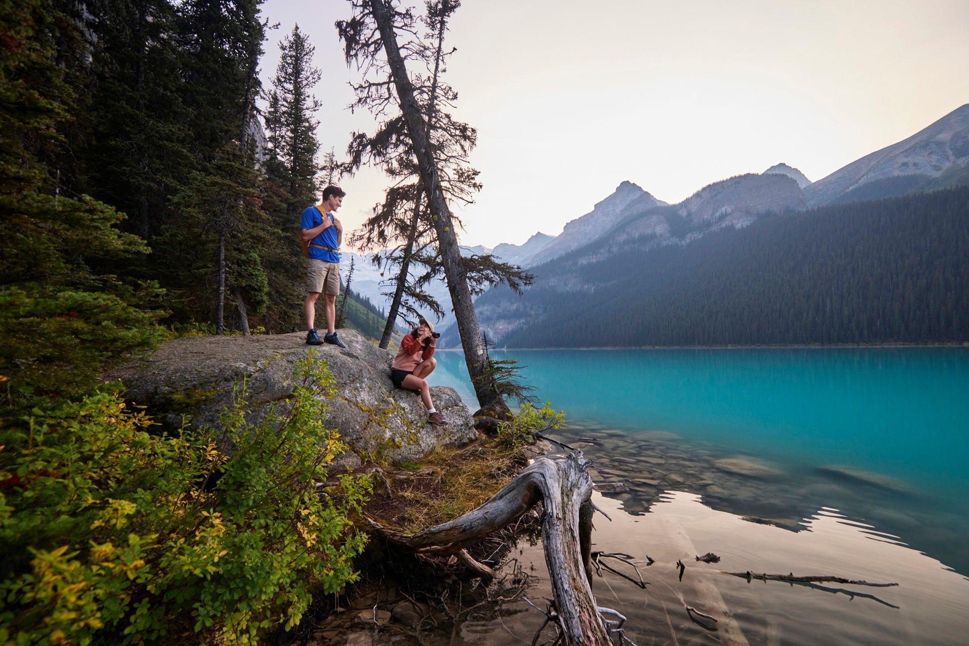 @Travel Alberta / Colin Way Two people near a turquoise lake, mountains in the background. Man standing, looking at phone. Woman sitting, taking photos.