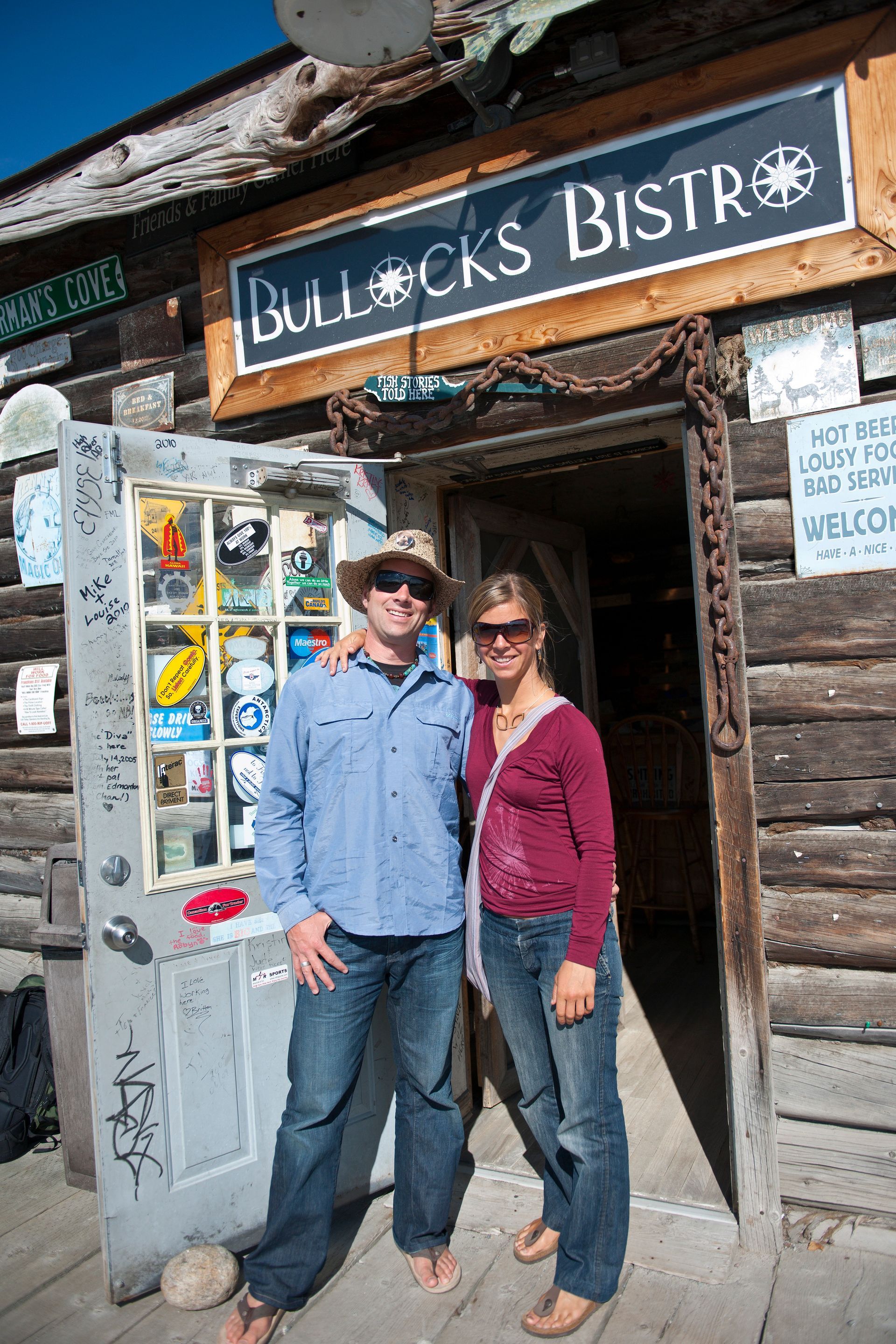 Twee mensen staan ​​lachend met zonnebrillen op buiten Bullocks Bistro, onder een houten uithangbord.