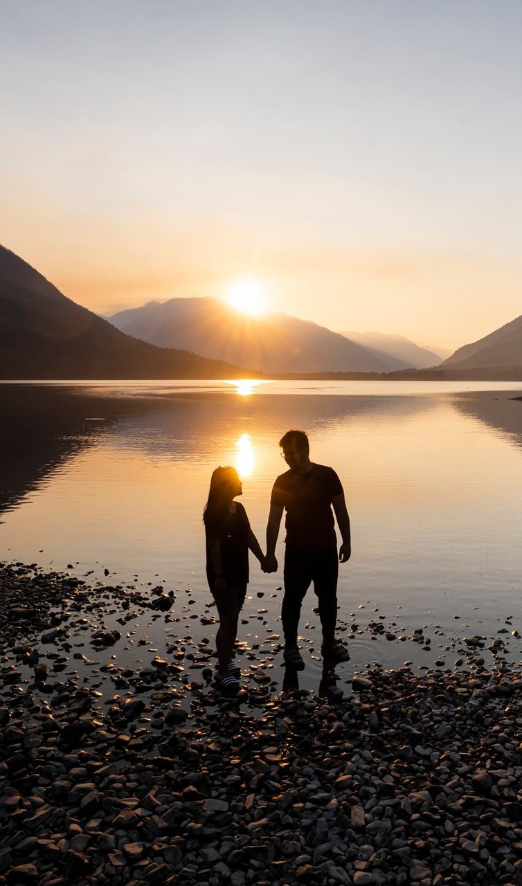 Twee mensen houden elkaars hand vast bij zonsondergang aan een meer, hun silhouetten steken af ​​tegen het kalme, spiegelende water en de bergen.