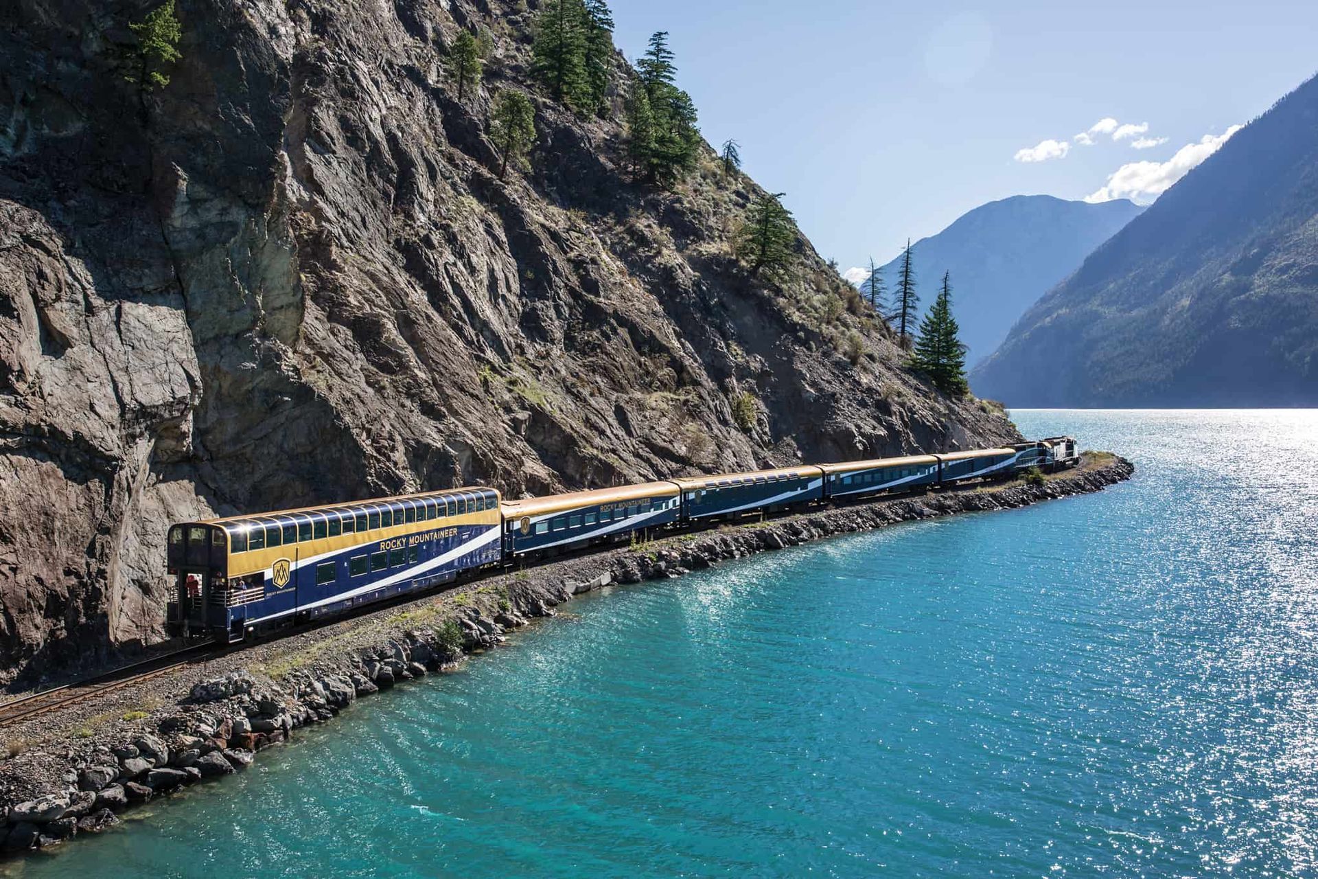 Train travels along a narrow track next to a turquoise lake and rocky mountain, under a sunny sky.