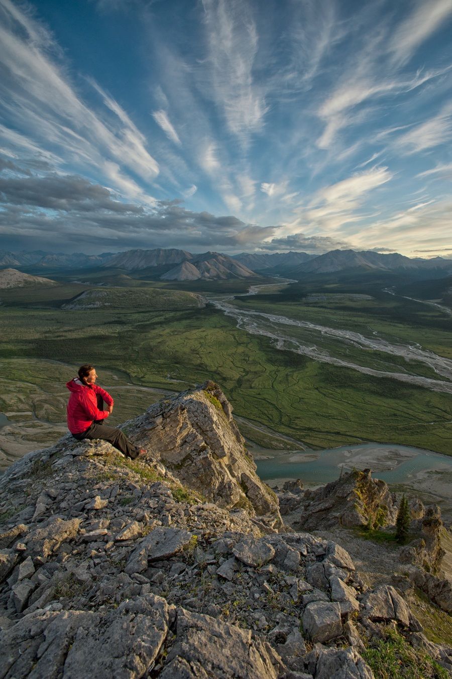 @matherpeter@peter.mather.photography Person in red jacket sits on rocky peak, looking at a valley and sky with streaky clouds.