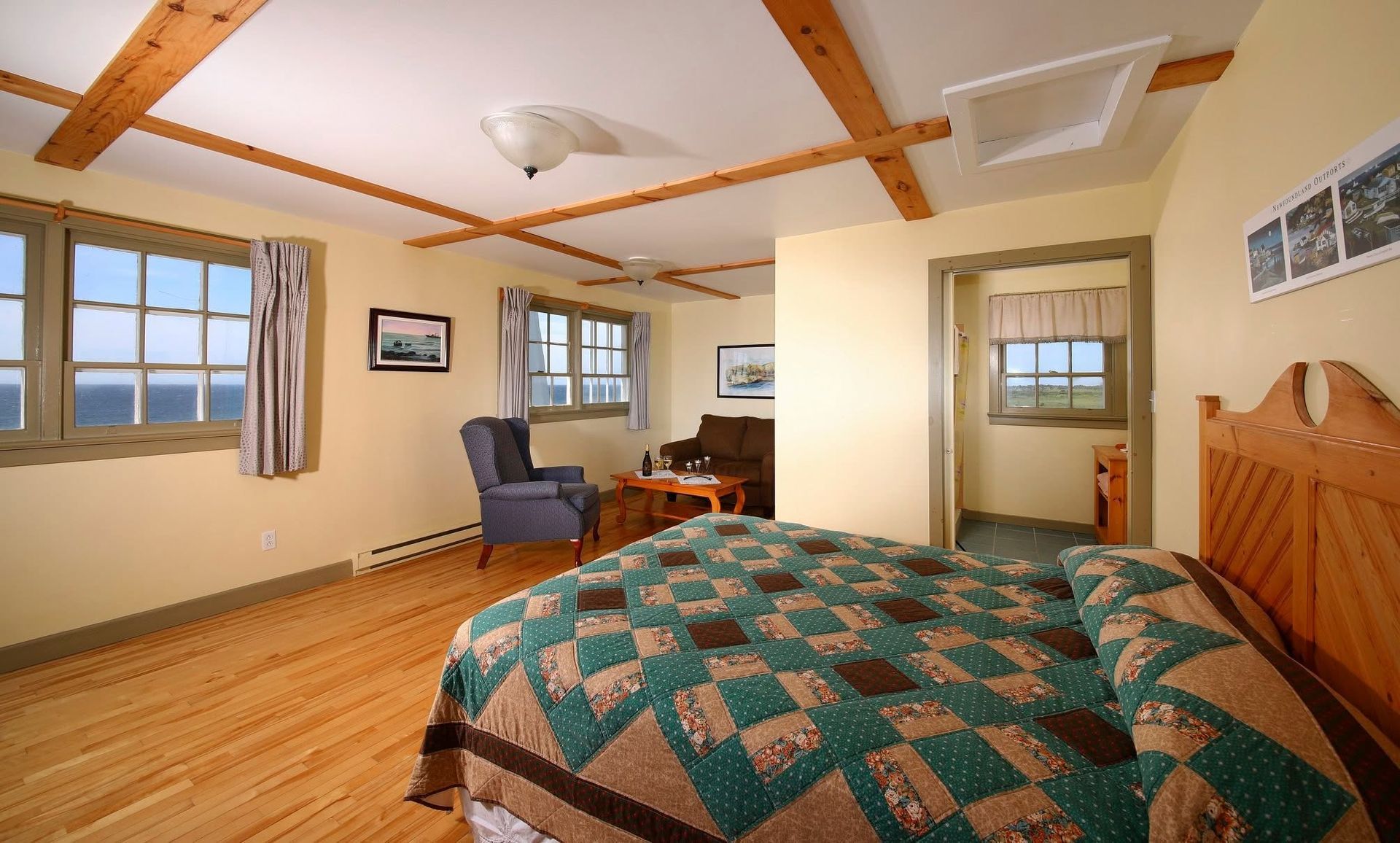 Lichte slaapkamer met gewatteerd bed, ramen, fauteuil, houten balken en een klein bureau.