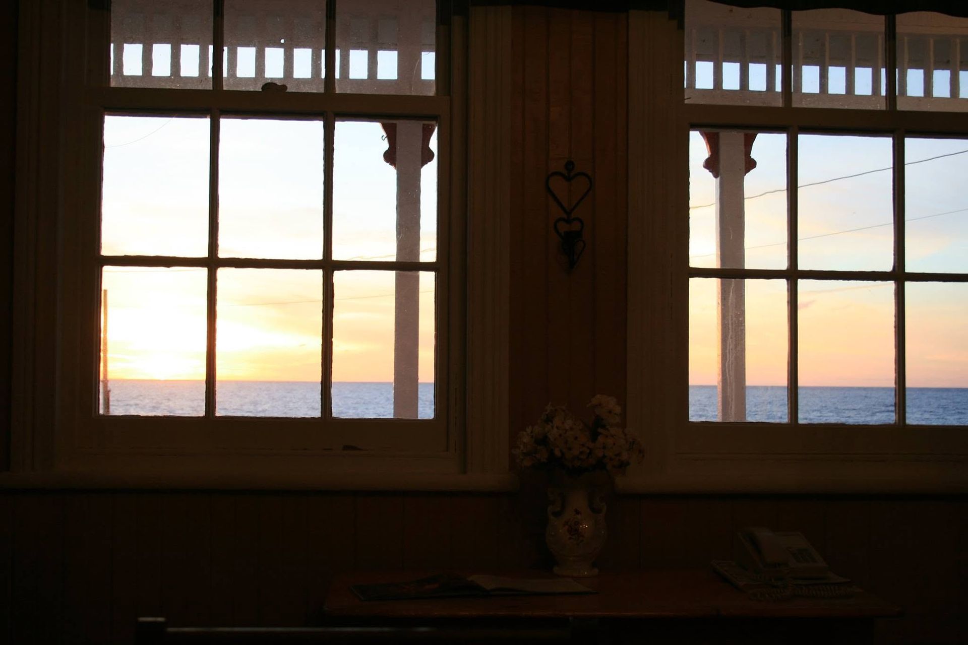 Zonsondergang gezien door twee ramen boven een tafel met een vaas bloemen in een kamer.
