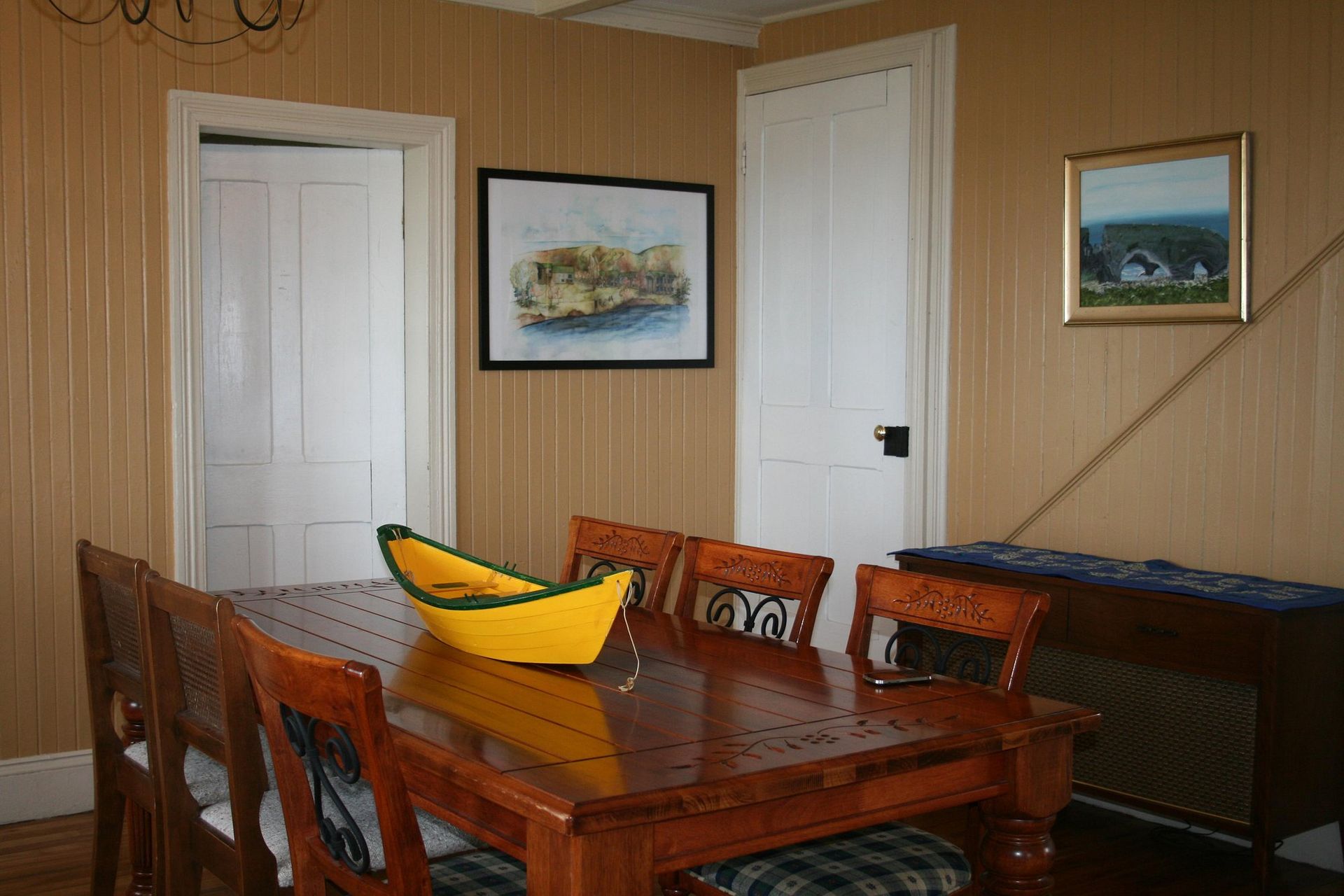 Eetkamer met houten tafel, bananenstuk in het midden en ingelijste schilderijen aan houten lambrisering.