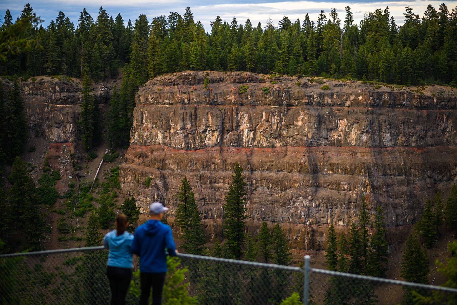 Twee mensen kijken uit over een beboste kloof in Chasm Provincial Park, Canada met hoge, gelaagde kliffen onder een bewolkte hemel.