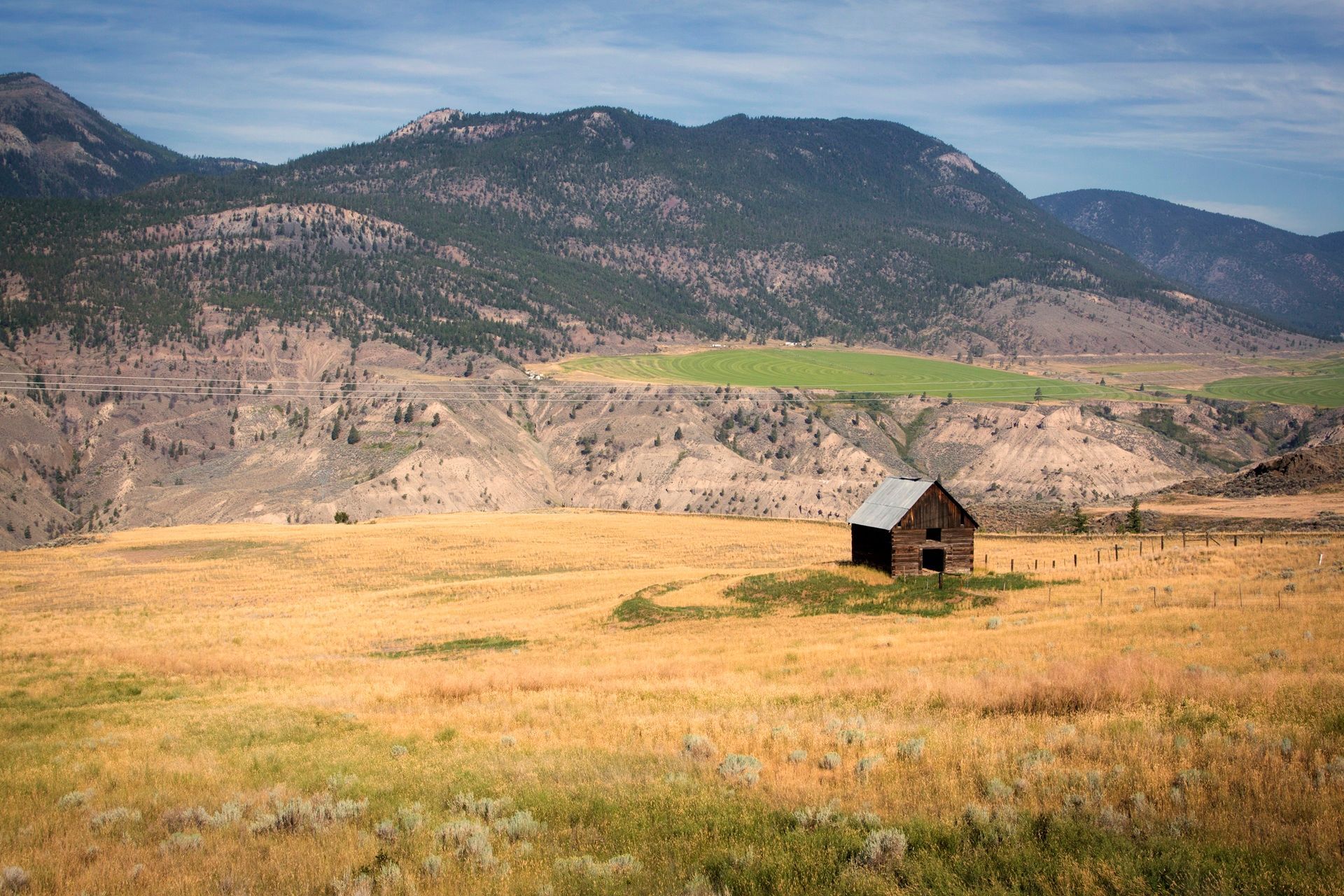 Een kleine houten hut in Canada in een droog, goudkleurig veld met bergen op de achtergrond onder een blauwe hemel.
