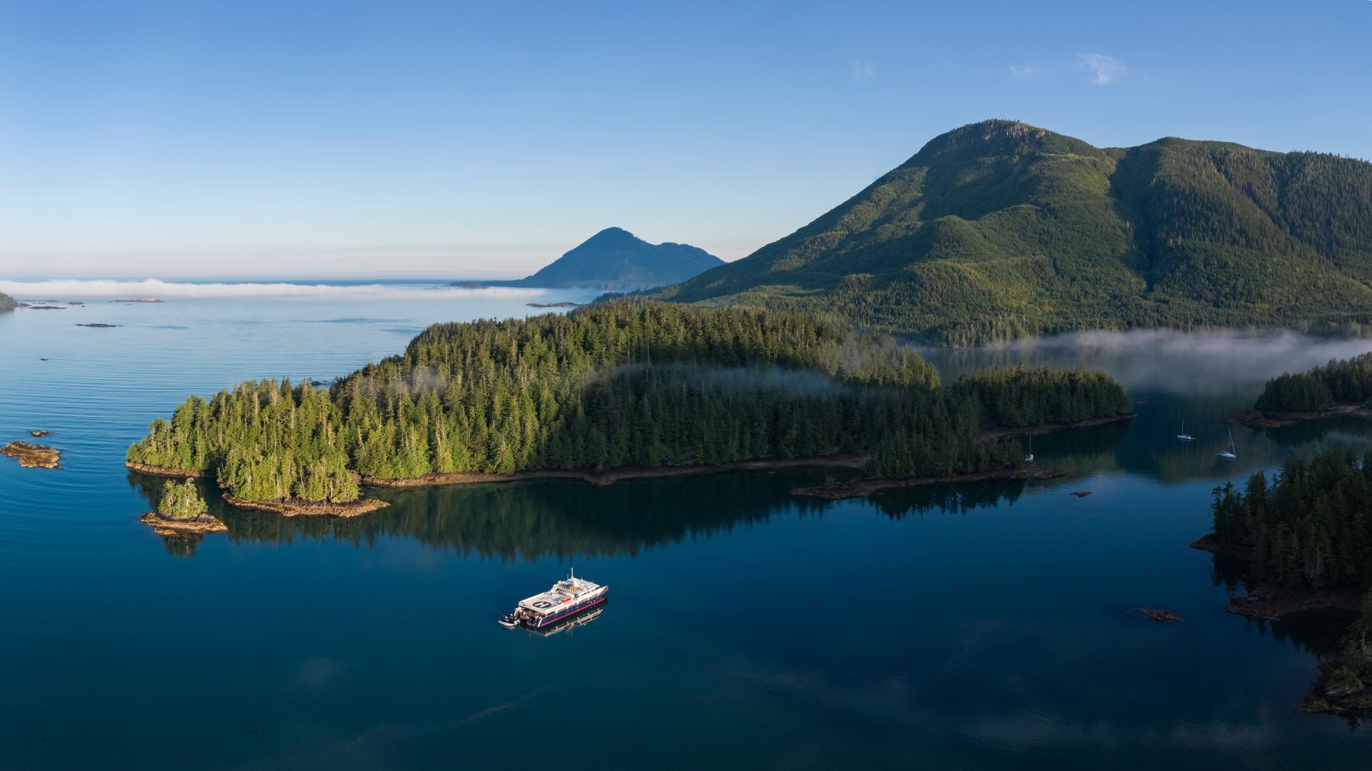Een groen eiland aan de kust van British Columbia met het schip Cascadia van Maple Leaf Adventures en mistige bergen op de achtergrond