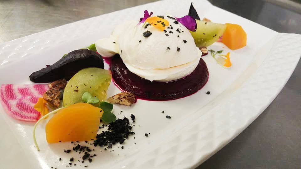 Een dessert op een wit bord, bestaande uit witte mousse, donkere bessensaus, schijfjes fruit en verkruimelde garnering.