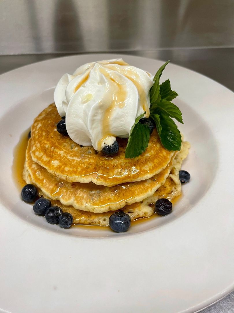 Een stapel pannenkoeken, bedekt met slagroom en munt, en gegarneerd met bosbessen, op een wit bord.