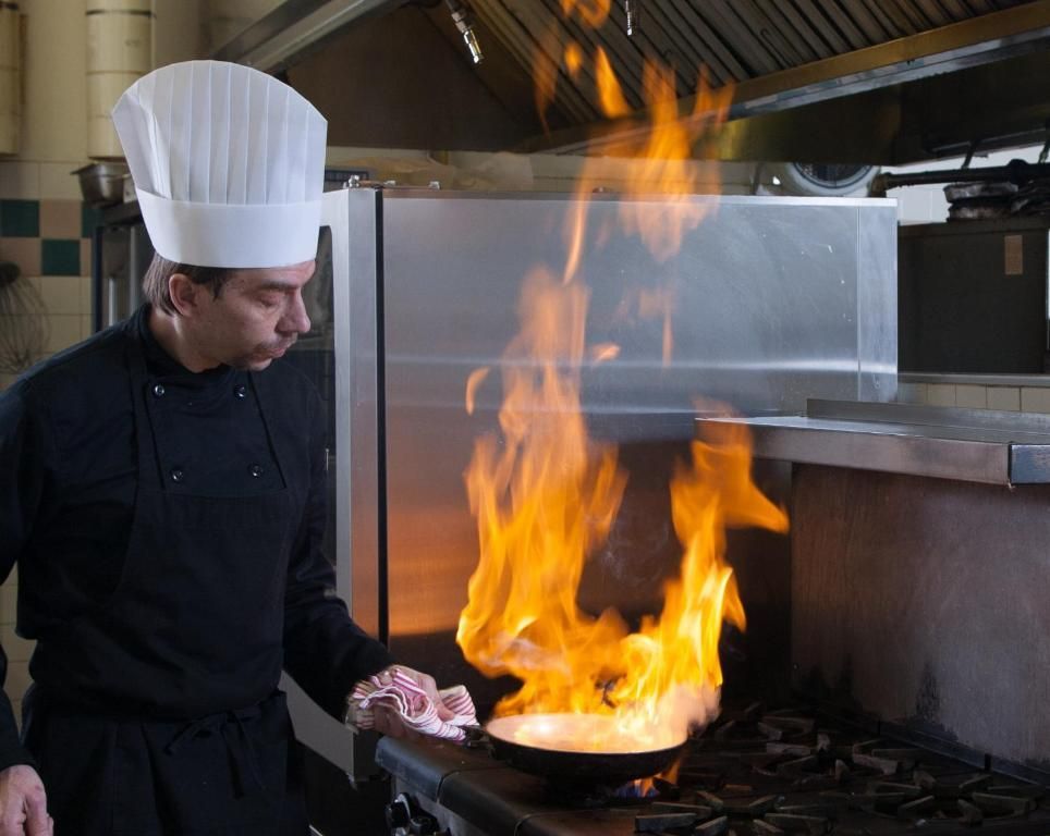 Een chef-kok met een witte koksmuts staat te koken aan een fornuis, terwijl een grote vlam uit een pan opstijgt in een professionele keuken.