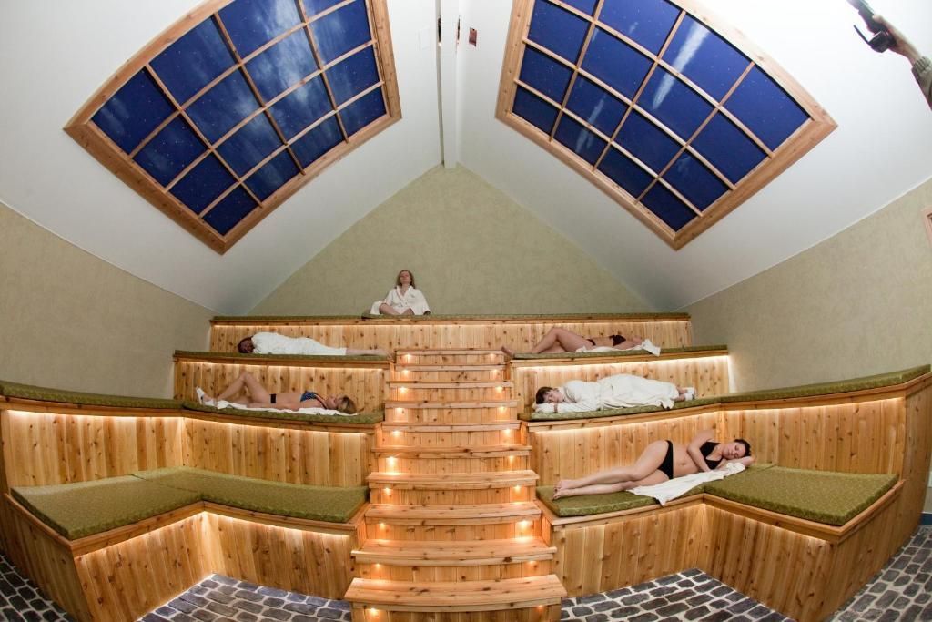 Saunaruimte met houten banken in verschillende niveaus, warme verlichting en mensen die onder dakramen ontspannen.