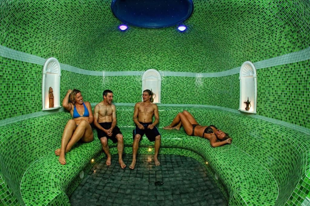 Vier mensen ontspannen in een groene mozaïeksauna met gewelfde nissen en blauwe plafondlampen.