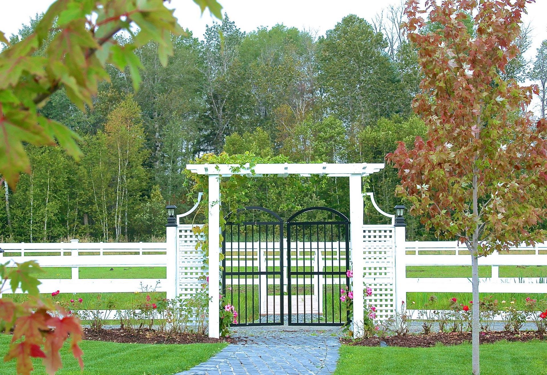 Witte tuinpoort met spijlen van traliewerk en zwarte ijzeren deuren, centraal gelegen in een omheinde tuin met bomen in de herfst.