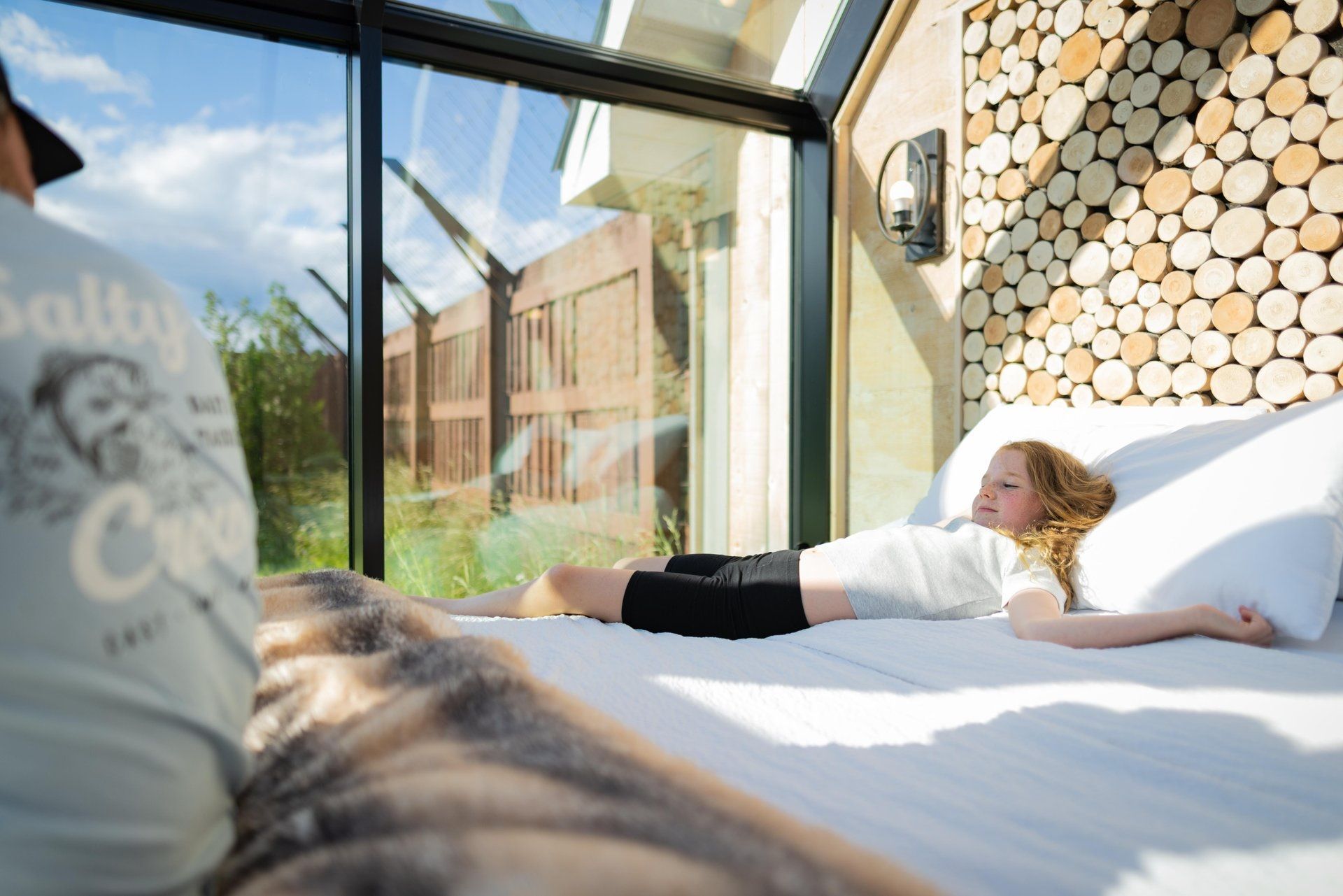 Een persoon ligt op een bed in een lichte kamer met glazen wanden en kijkt uit op een tuin.