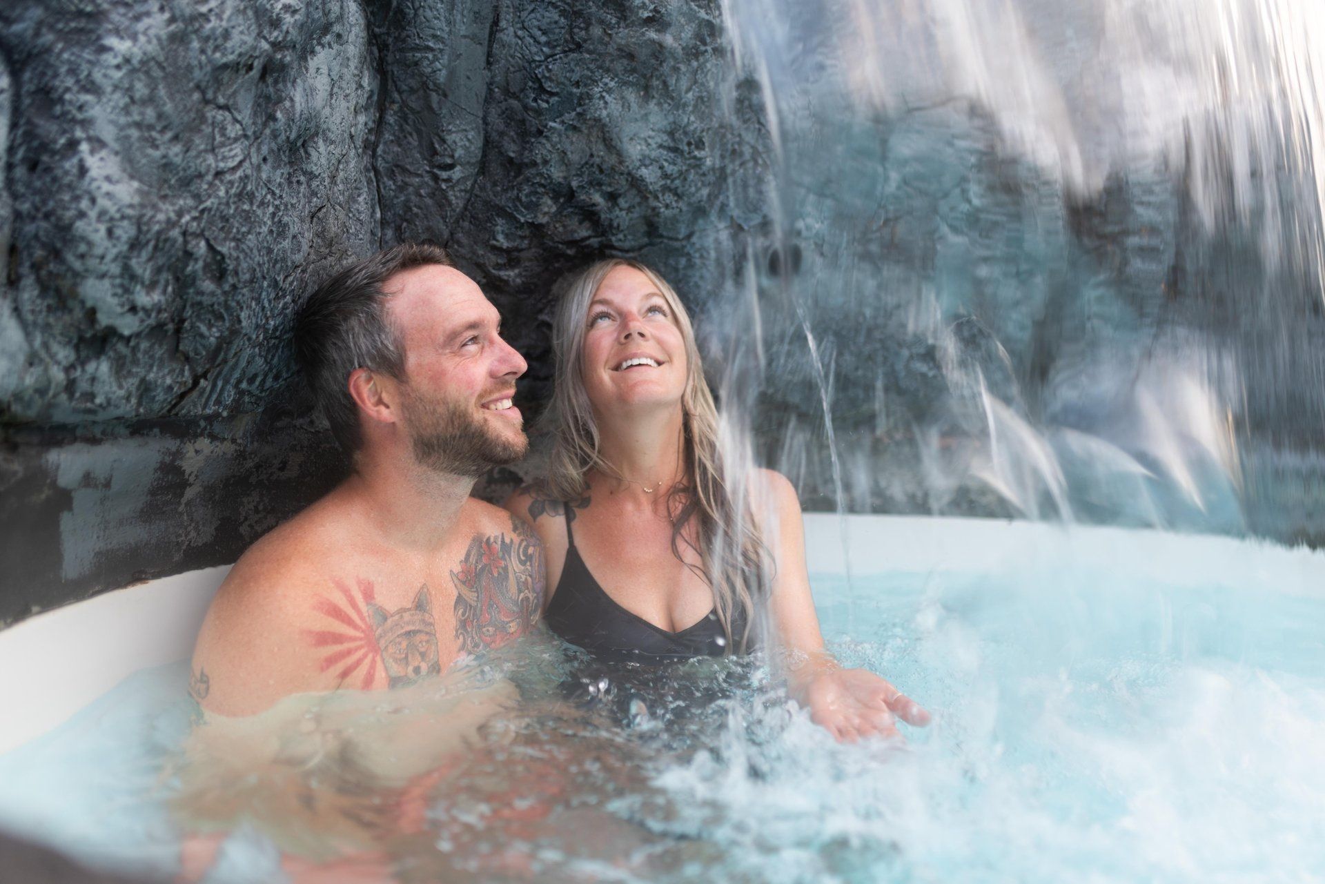 Twee mensen ontspannen in een stomend heet bad naast een waterval, glimlachend en omhoogkijkend.
