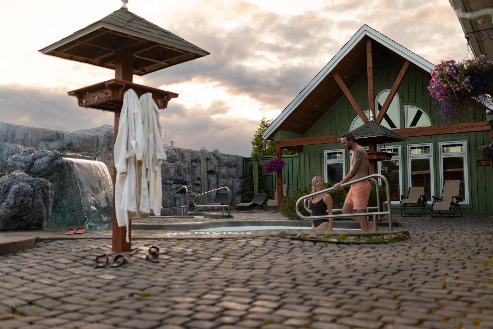 Buitenpatio van het resort met een rieten parasol, ligstoelen en een gebouw met een groen dak bij zonsondergang.