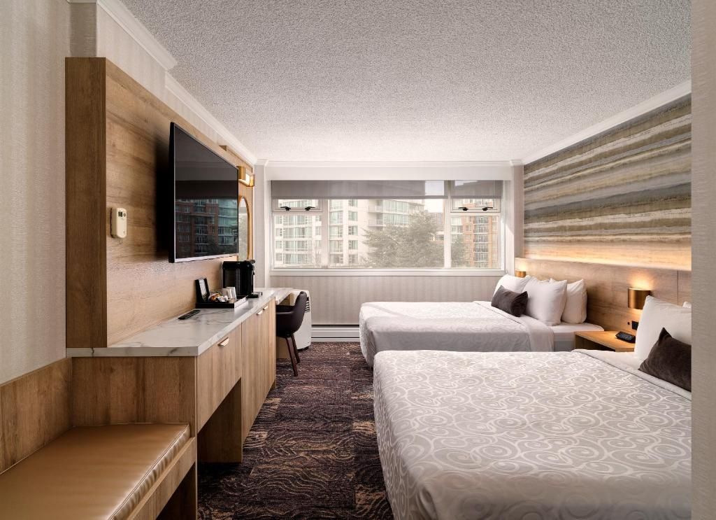 Moderne hotelkamer met twee bedden, aan de muur gemonteerde tv, bureau en een groot raam met uitzicht op de stadsgebouwen.