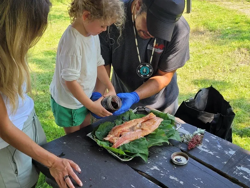 Mensen bereiden een vis voor op groene bladeren aan een picknicktafel in de buitenlucht, met handschoenen en keukengerei binnen handbereik.