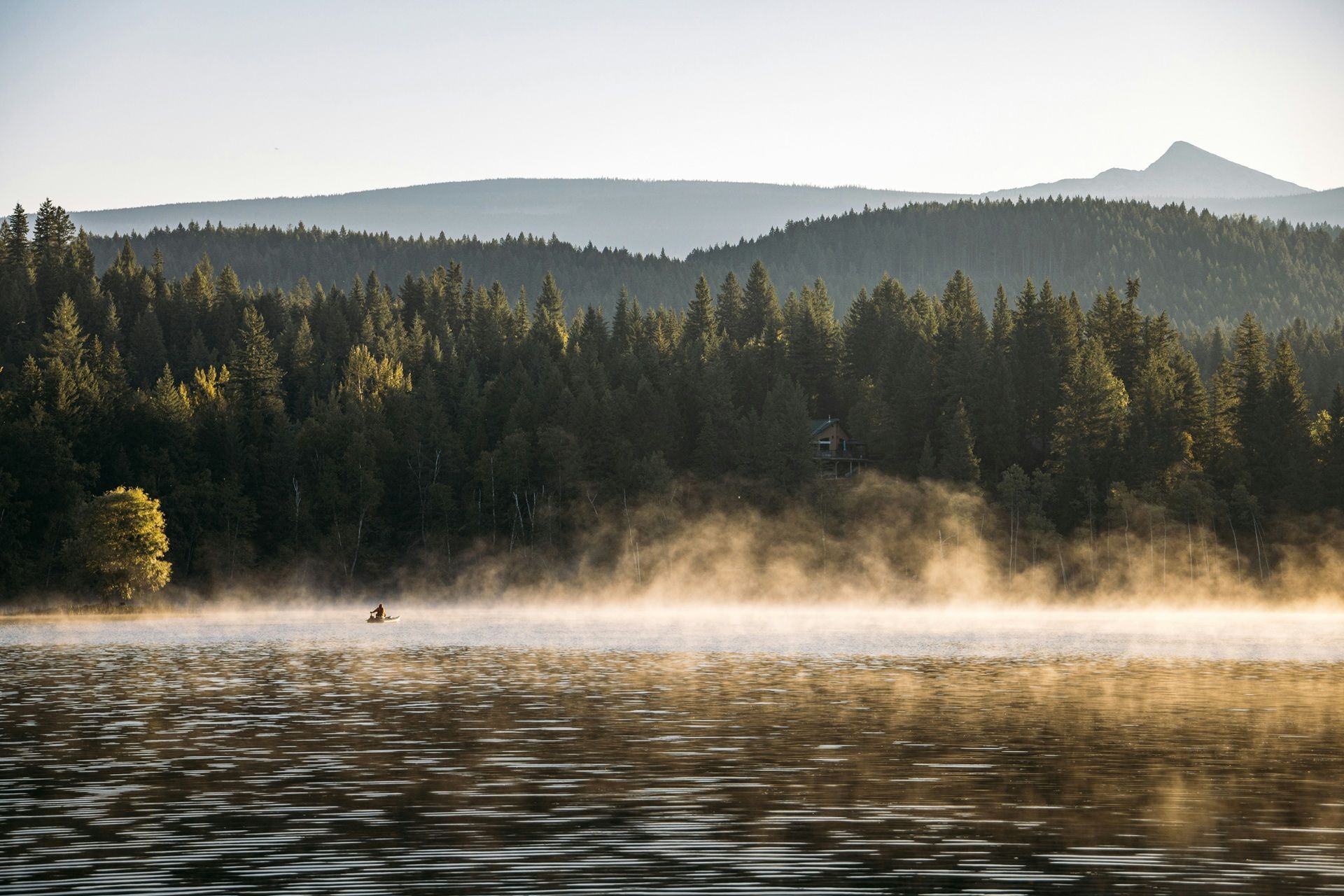 Mist stijgt op boven Clear Water Lake in Wells Gray Provincial Park met een beboste oever en wazige bergen bij zonsopgang.