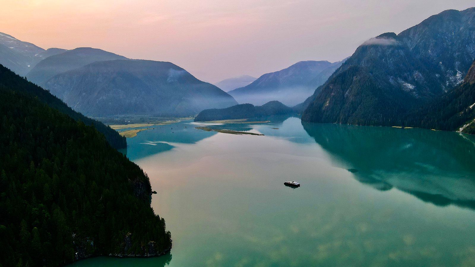 Turkooizen zee in British Columbia tussen donkere bergen bij zonsopkomst, met het schip Cascadia van Maple Leaf Adventures op het kalme water.