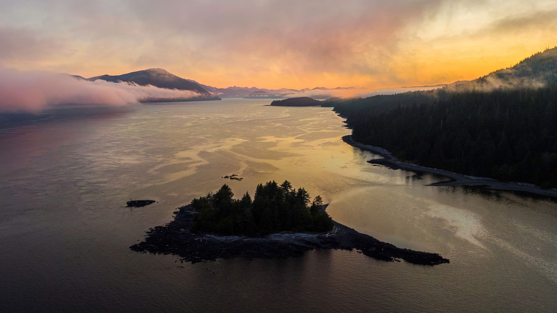 Zonsondergang boven Haida Gwaii, British Columbia, met een smal eiland met donkere bomen, weerspiegeld in kalm water met mistige bergen op de achtergrond