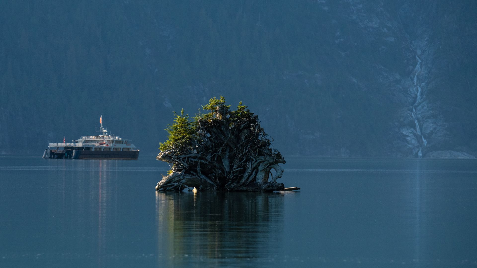 Het schip Cascadia van Maple Leaf Adventures bij een piepklein bebost eilandje voor de kust van Britsh Columbia op kalm blauw water in de schemering