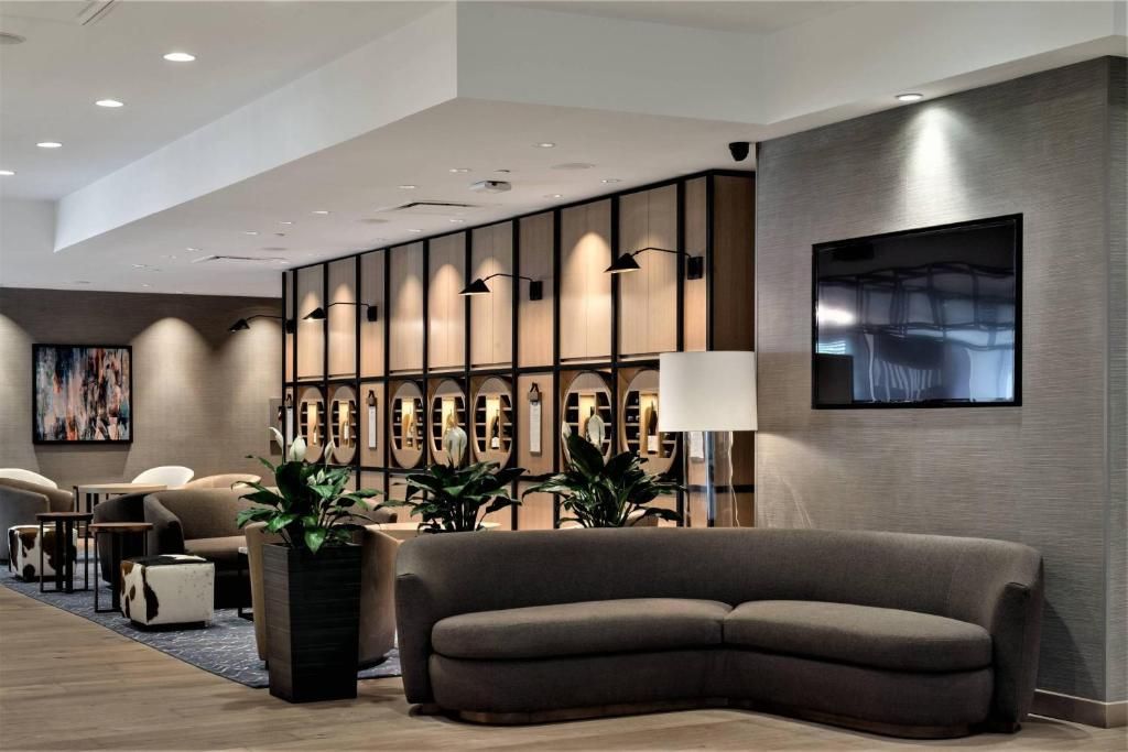 Moderne hotellobby met gebogen grijze bank, aan de muur gemonteerde tv en decoratieve planken in neutrale tinten.