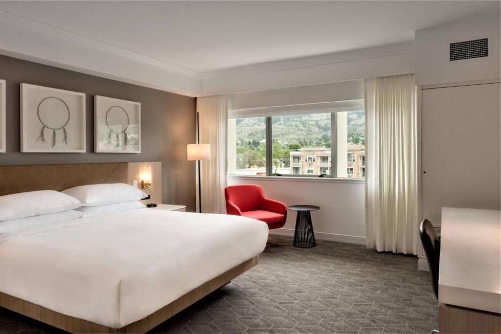 Moderne hotelkamer met een kingsize bed, rode stoel, bureau en een groot raam met uitzicht op de bomen.