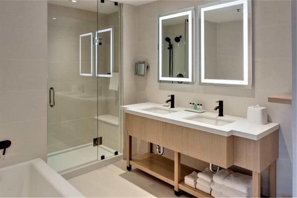 Moderne badkamer met glazen douchecabine, dubbele wastafel en verlichte spiegels in neutrale tinten.