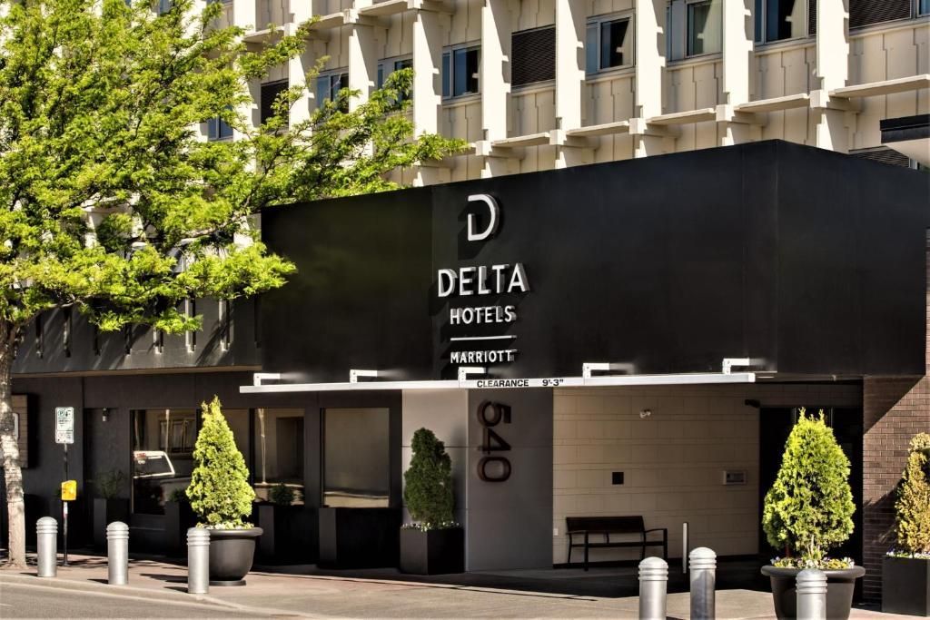 De ingang van Delta Hotels met een zwart zonnescherm, potplanten en de gevel van het gebouw bij daglicht.