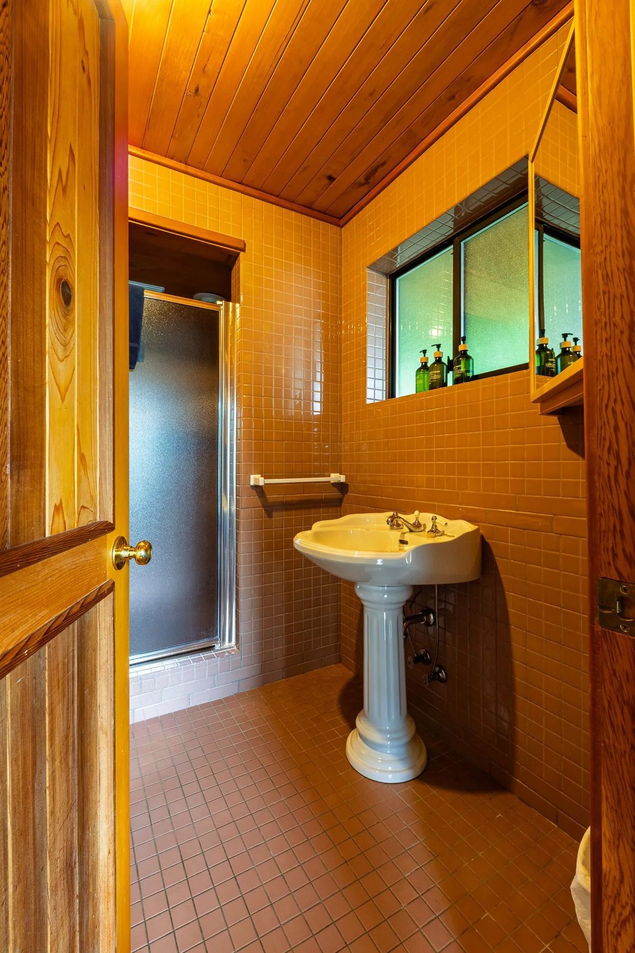 Kleine badkamer met houten lambrisering, wastafel op een voetstuk, betegelde douche en vensterbank.
