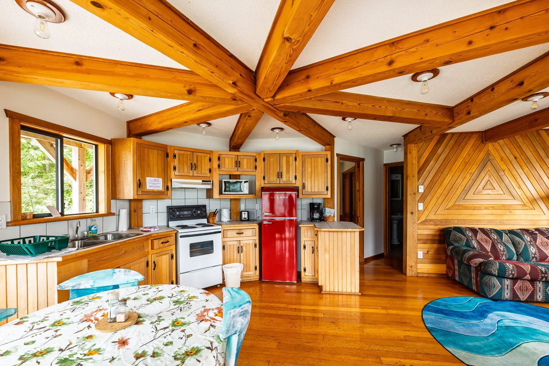 Lichte, rustieke keuken-woonkamer met houten balken, rode koelkast en bloemenpatroon op het tafelkleed.