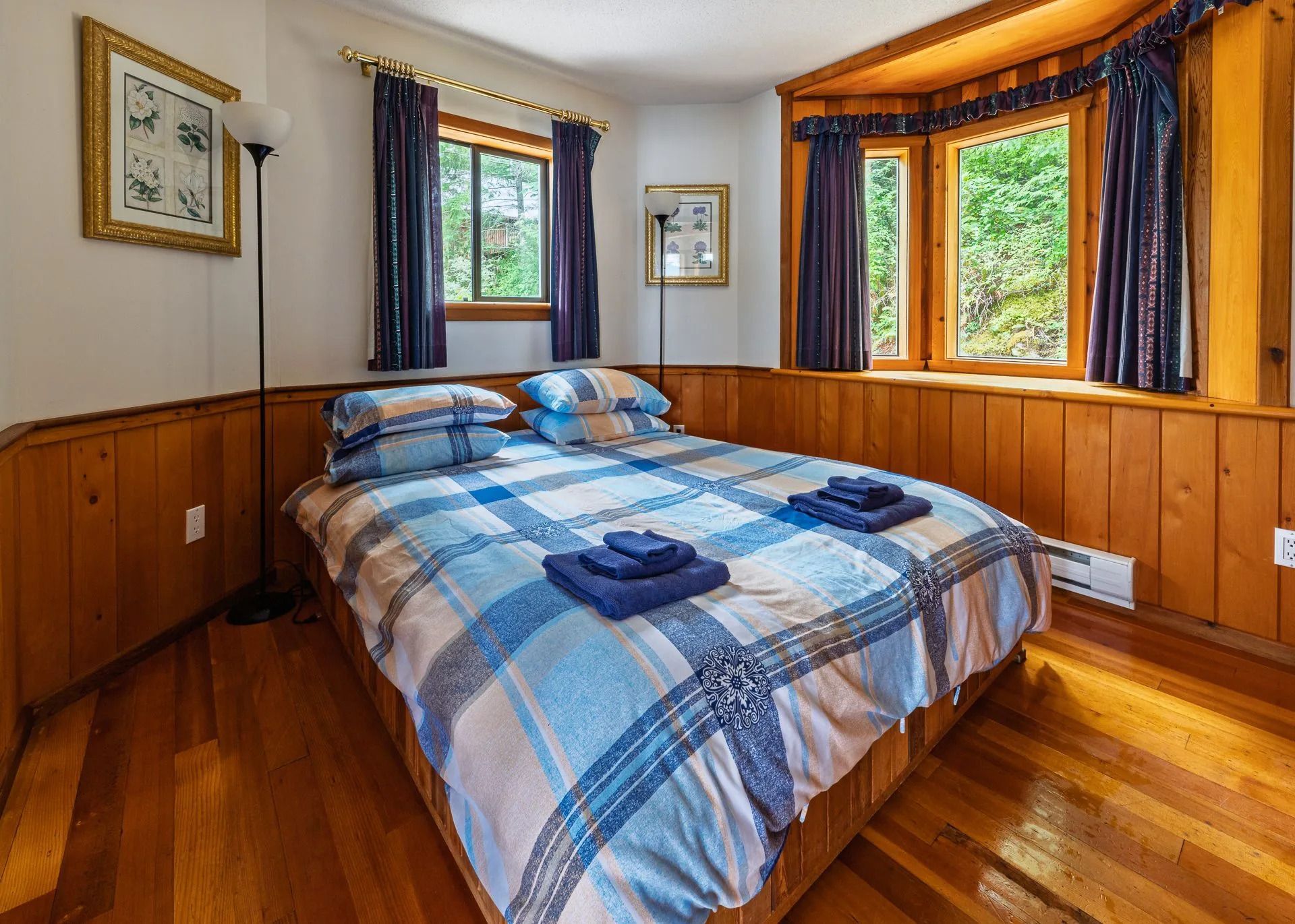 Gezellige slaapkamer met een blauw geruite sprei, houten lambrisering en ramen met uitzicht op het groen.