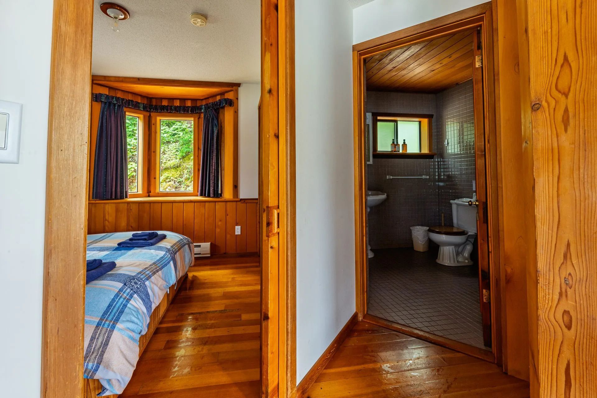 Uitzicht vanuit de gang op een slaapkamer met houten lambrisering en een aangrenzende badkamer met blauw beddengoed en een betegelde douche.