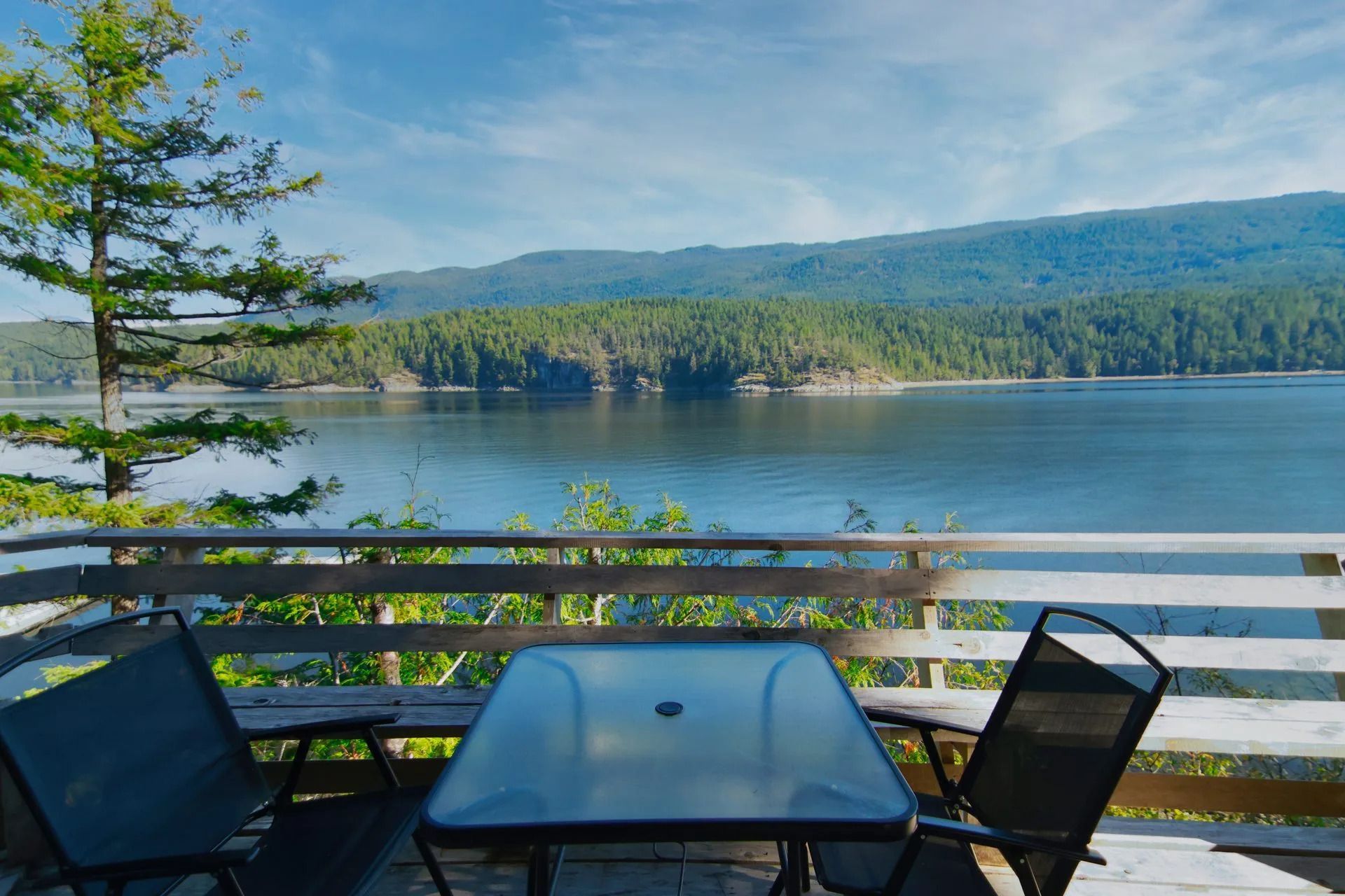 Terrastafel en stoelen met uitzicht op een rustig meer en beboste bergen op een zonnige dag.