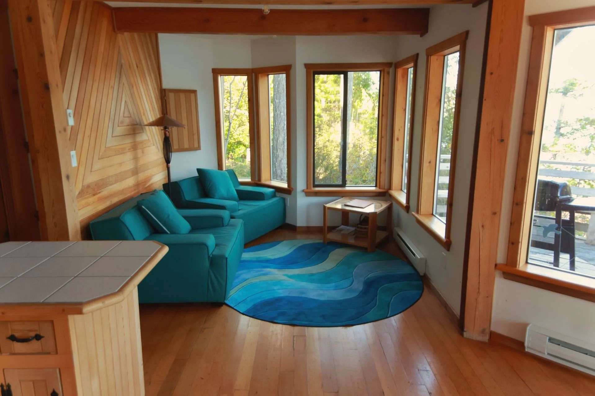Gezellige woonkamer met turquoise bank, blauw vloerkleed, houten balken en grote ramen met uitzicht op een terras.