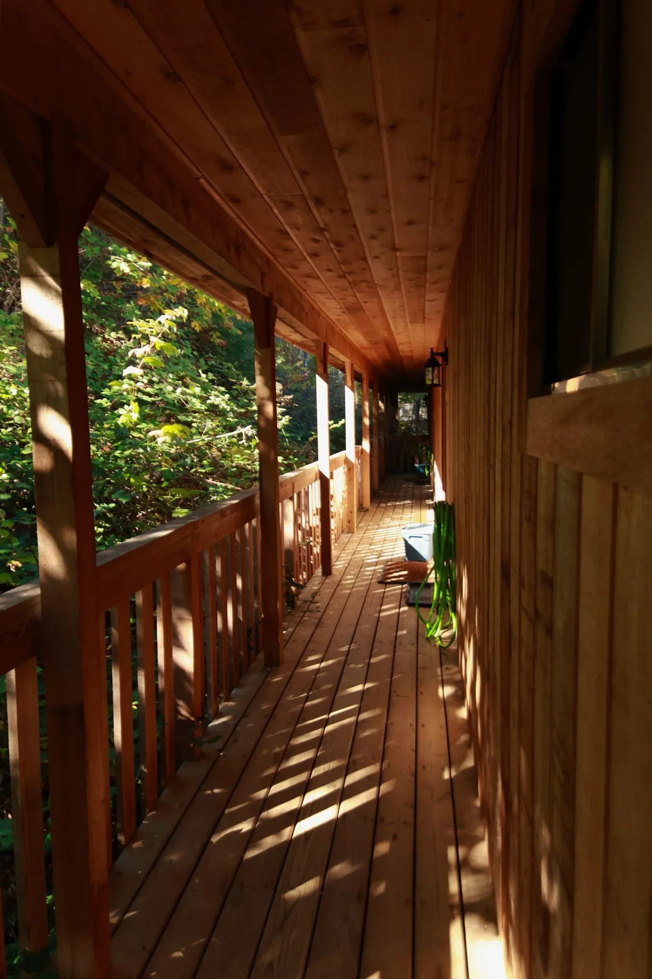 Een zonnige houten veranda met balustrade, potplant en uitzicht op het bos dat lange schaduwen werpt.