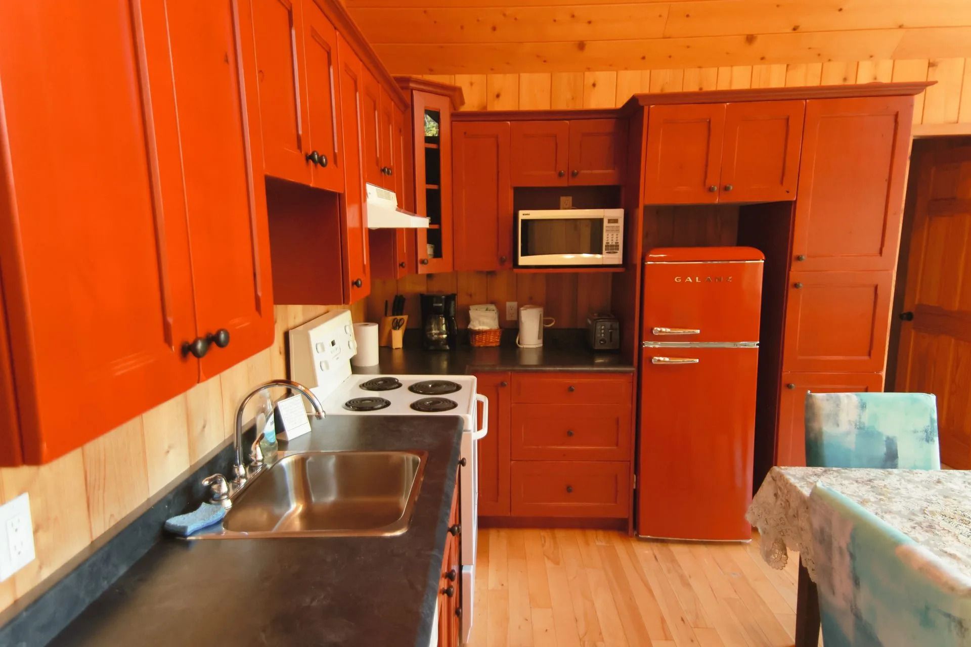 Houten keuken met oranje kastjes, wit fornuis, rode koelkast en een tafel bij het raam.