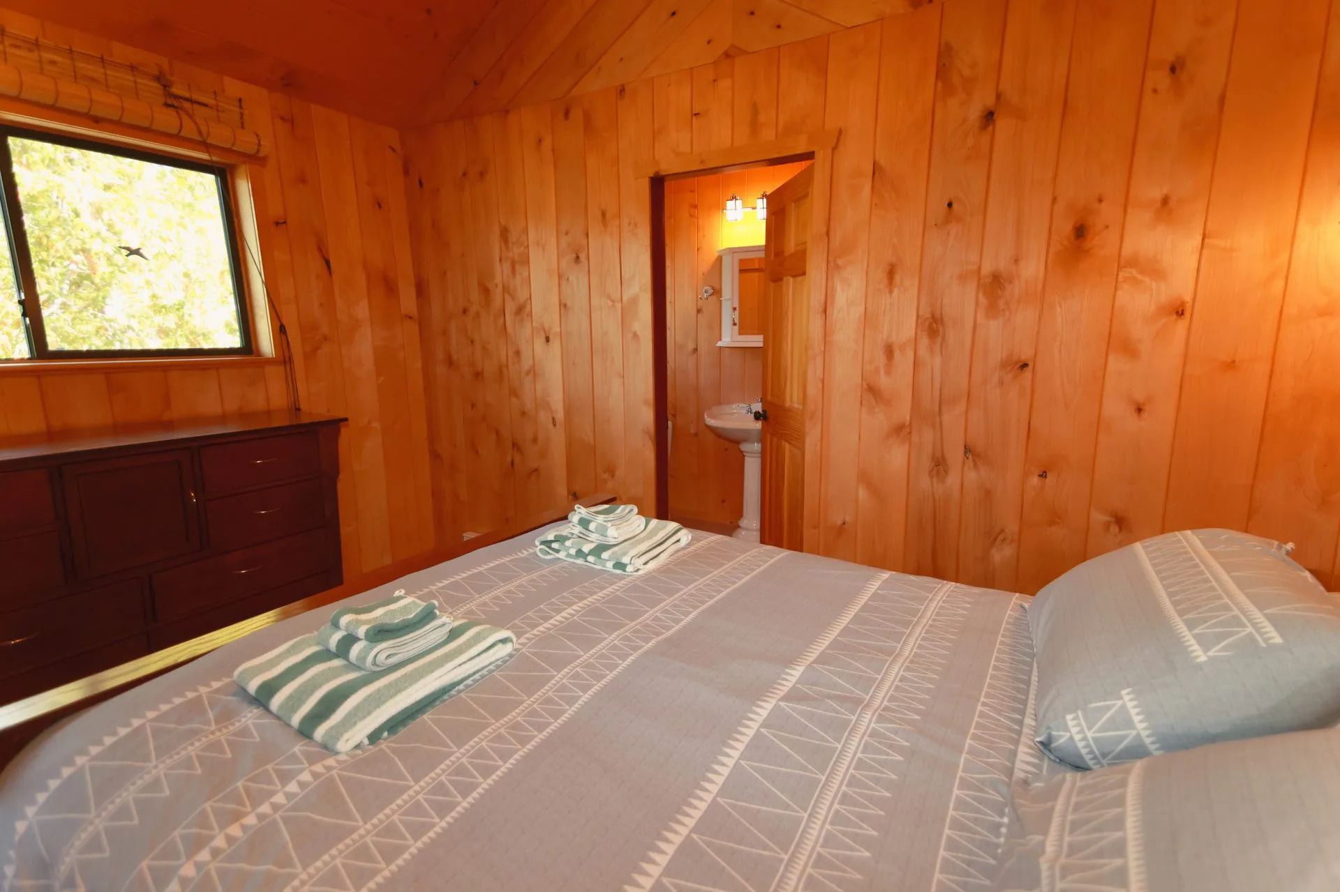 Slaapkamer met houten lambrisering, een bed, gestreepte kussens en een klein raam boven een dressoir.