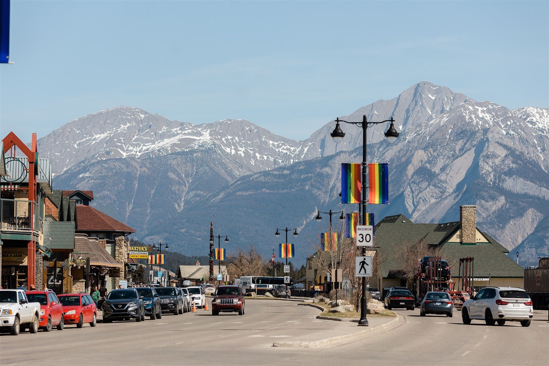 Een straat in het centrum van Jasper in Canada met auto's, kleurrijke spandoeken en besneeuwde bergen onder een helderblauwe hemel.