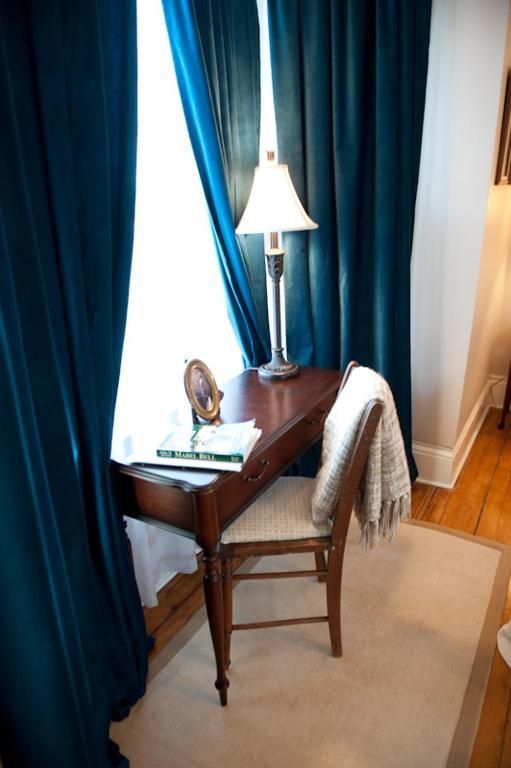 Een klein houten bureau bij blauwe gordijnen met een lamp, een ingelijste foto en een gedrapeerde sjaal in een zonnige kamer.