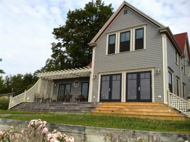 Grijze woning met twee verdiepingen, een veranda aan de voorzijde, grote ramen en een stenen keermuur op een heuvel.